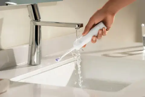 Mano che sciacqua uno spazzolino elettrico sotto un rubinetto. Ideale per chi soffre di gengive ritirate.